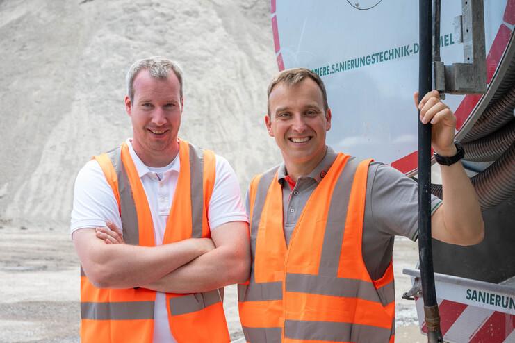 Zwei Männer in orangefarbener Warnkleidung stehen vor einer Baustelle mit Sand- und Kieshaufen im Hintergrund.