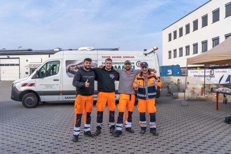 Vier Mitarbeiter in orangefarbener Arbeitskleidung stehen lächelnd vor einem Firmenfahrzeug auf einem Betriebshof.